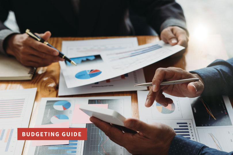 Two individuals sitting at a desk with their hands holding a calculator, pens, and budget graphs